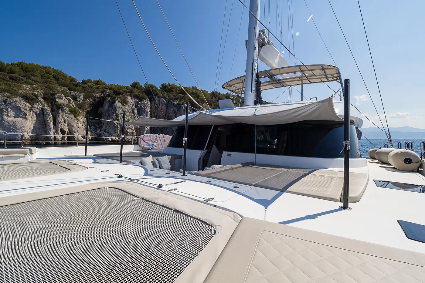 The deck of the Sunreef 50 yacht, featuring spacious sunbathing areas with netted trampolines, cushioned seating, and shaded lounging spots, with scenic cliffs in the background.