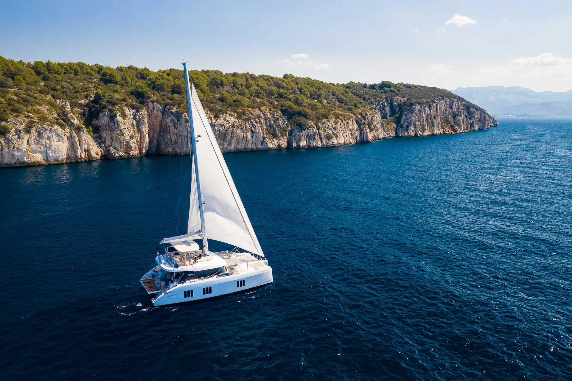 The Sunreef 50 yacht gracefully sailing along a rugged coastline, with its sails fully extended against a backdrop of clear blue skies and deep blue waters.