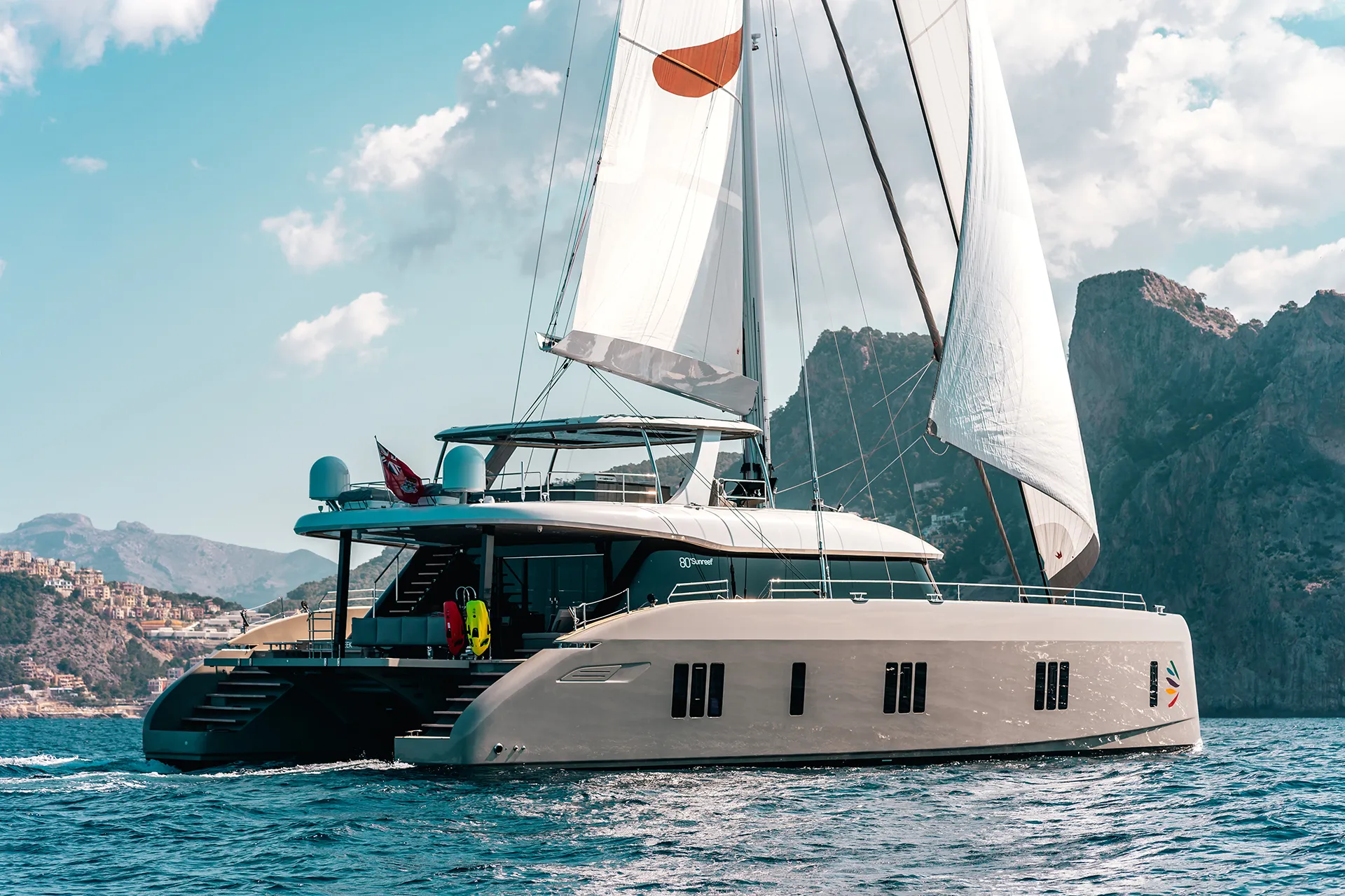 Sunreef 80 Katamaran unter vollen Segeln, das Logo auf dem Segel hebt sich farbenfroh vom blauen Himmel ab, während die Yacht durch ruhiges Wasser segelt.